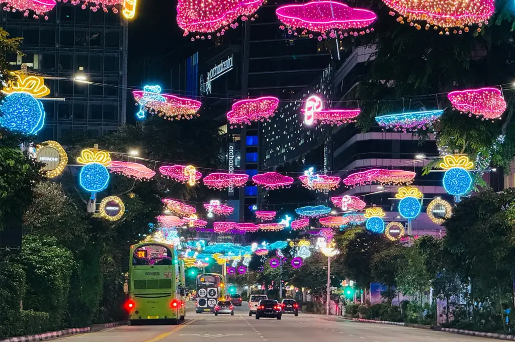 A photo of nighttime at Orchard Road, Singapore, with festive neon pink and blue cloud and gift lights over a busy street. A green double-decker bus and cars drive past lush trees and glowing city buildings after dark.