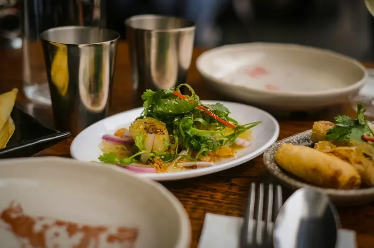 Table setting with asian dishes including fresh herb salad, spring rolls, and metal cups, arranged on a wooden table in a restaurant.