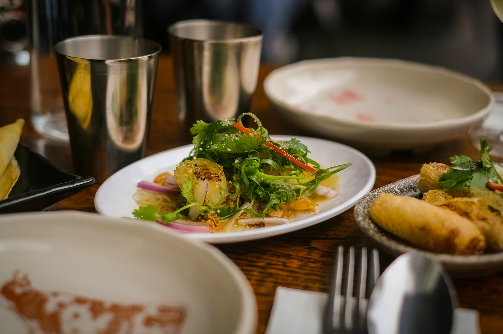 Table setting with asian dishes including fresh herb salad, spring rolls, and metal cups, arranged on a wooden table in a restaurant.