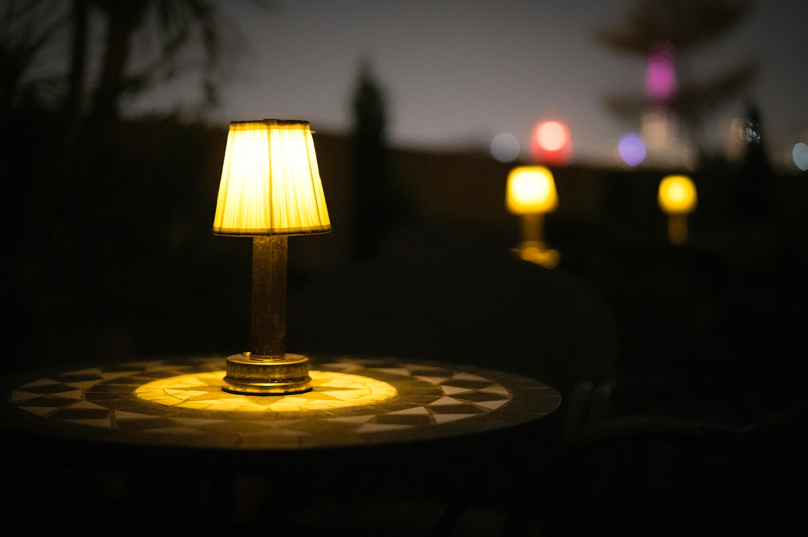 Warm glowing table lamp on a small café table at night, creating a cozy ambiance with soft blurred lights in the background.