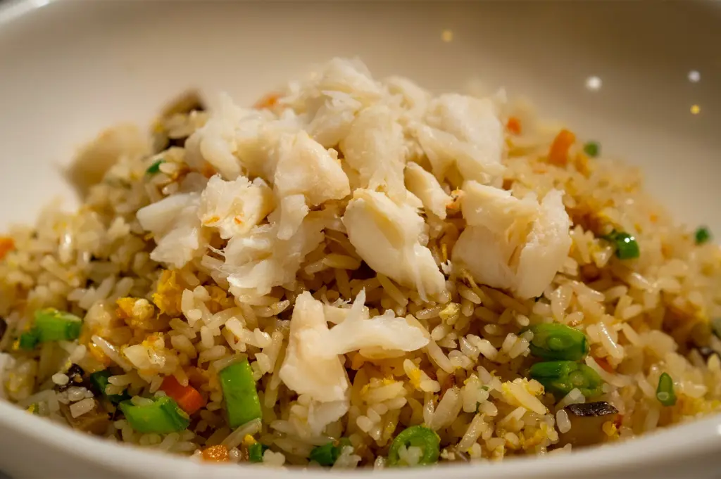 Close-up of crab fried rice topped with chunks of white crab meat, mixed with egg, green onions, and vegetables in a white bowl.