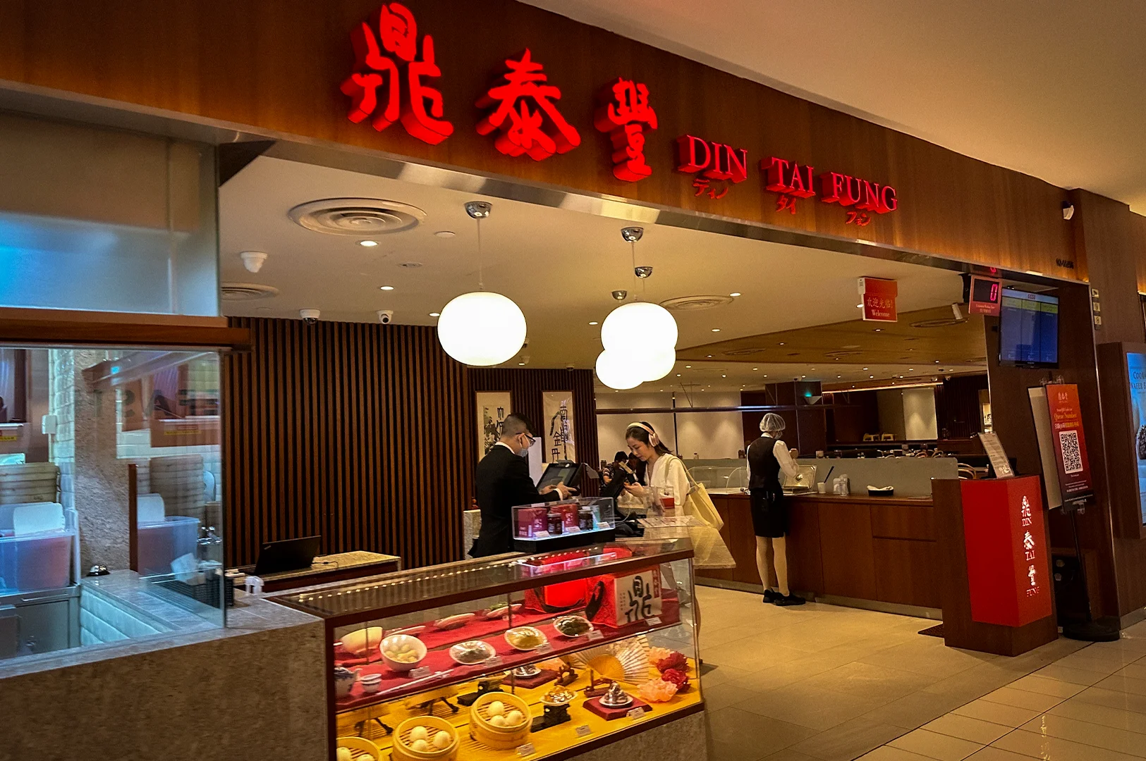 Front entrance of Din Tai Fung Singapore restaurant featuring illuminated signage, modern wooden interiors, and a display counter with signature dim sum dishes and xiao long bao models