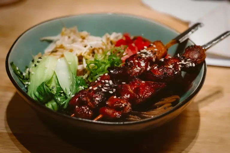 Bowl of noodles topped with grilled skewered meat, bok choy, bean sprouts, and sesame seeds in an Asian-style dish on a wooden table.