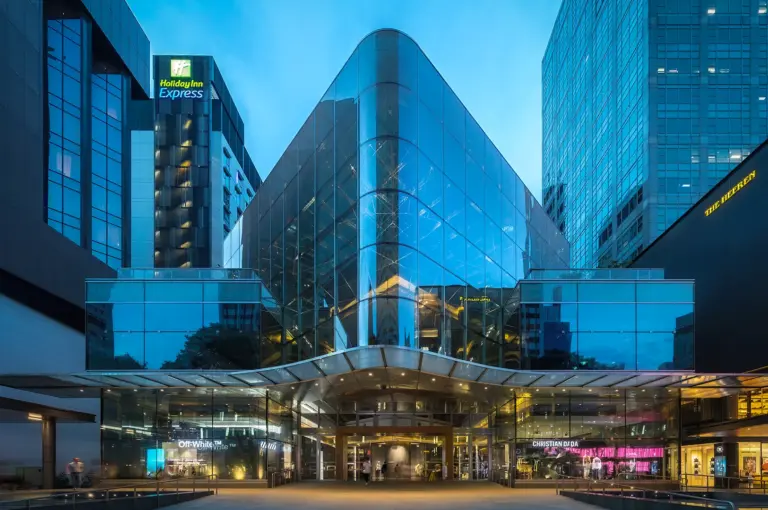 Illuminated glass facade of 270 Orchard Road (formerly Knightsbridge mall) in Singapore during blue hour. The striking architectural design features geometric glass panels, reflecting the city lights near the Holiday Inn Express and The Heeren.