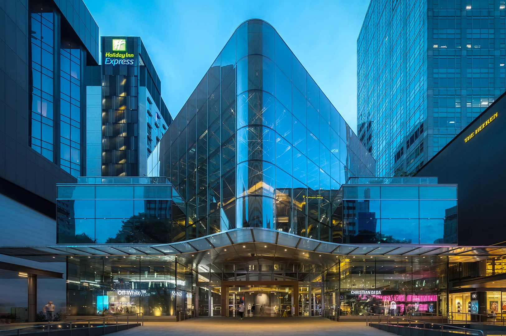 Illuminated glass facade of 270 Orchard Road (formerly Knightsbridge mall) in Singapore during blue hour. The striking architectural design features geometric glass panels, reflecting the city lights near the Holiday Inn Express and The Heeren.