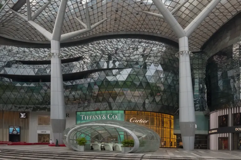 Exterior of ION Orchard in Singapore featuring a curved glass façade, lattice canopy structure, and luxury brand storefronts beneath a tall modern tower.