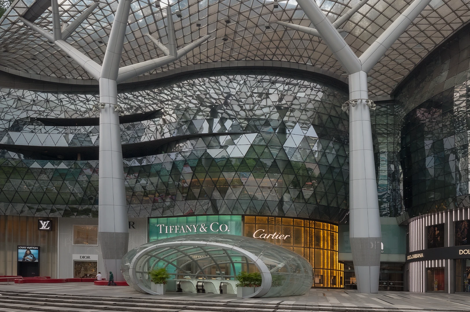 Exterior of ION Orchard in Singapore featuring a curved glass façade, lattice canopy structure, and luxury brand storefronts beneath a tall modern tower.