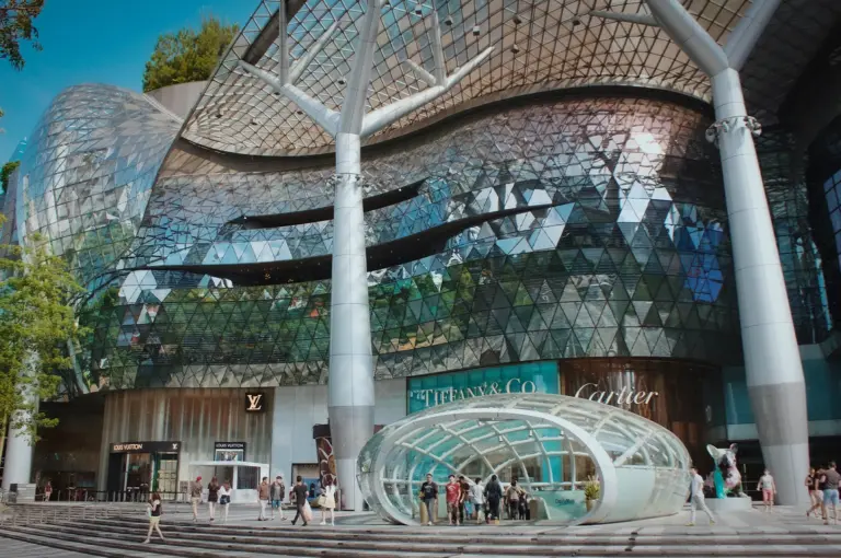 Exterior of ION Orchard shopping mall in Singapore with a geometric glass facade, luxury brand storefronts, and people walking near the entrance.