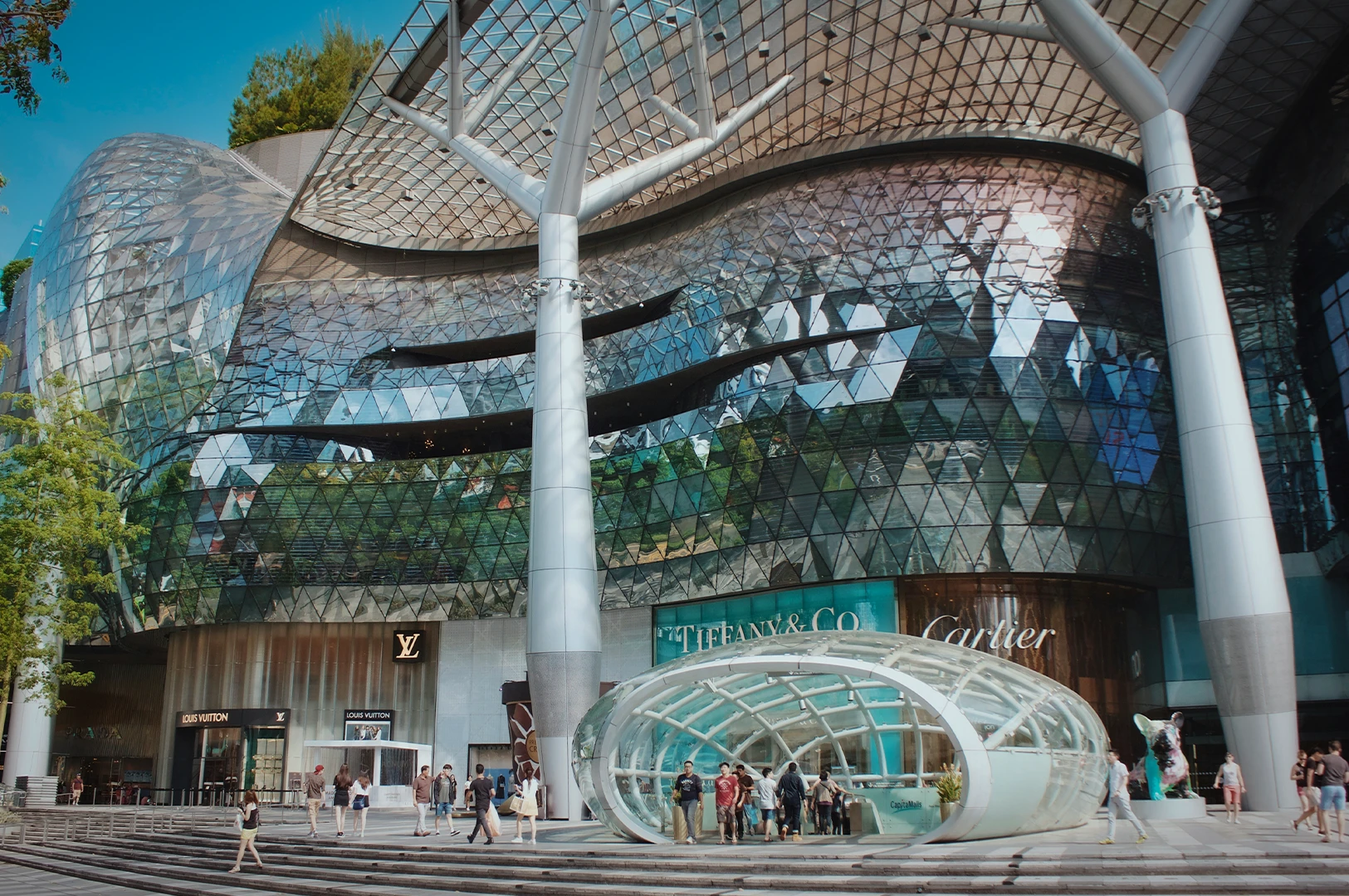 Exterior of ION Orchard shopping mall in Singapore with a geometric glass facade, luxury brand storefronts, and people walking near the entrance.