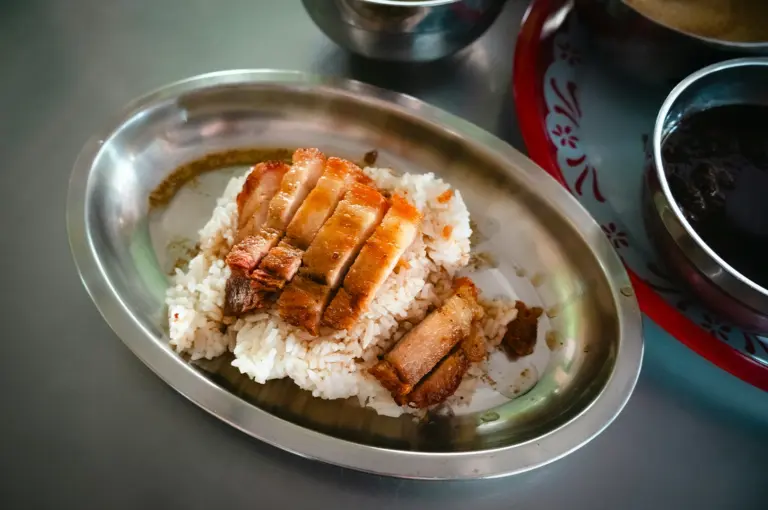 Plate of roasted pork slices served over white rice on a metal tray, with sauce and side dishes in a casual dining setting.