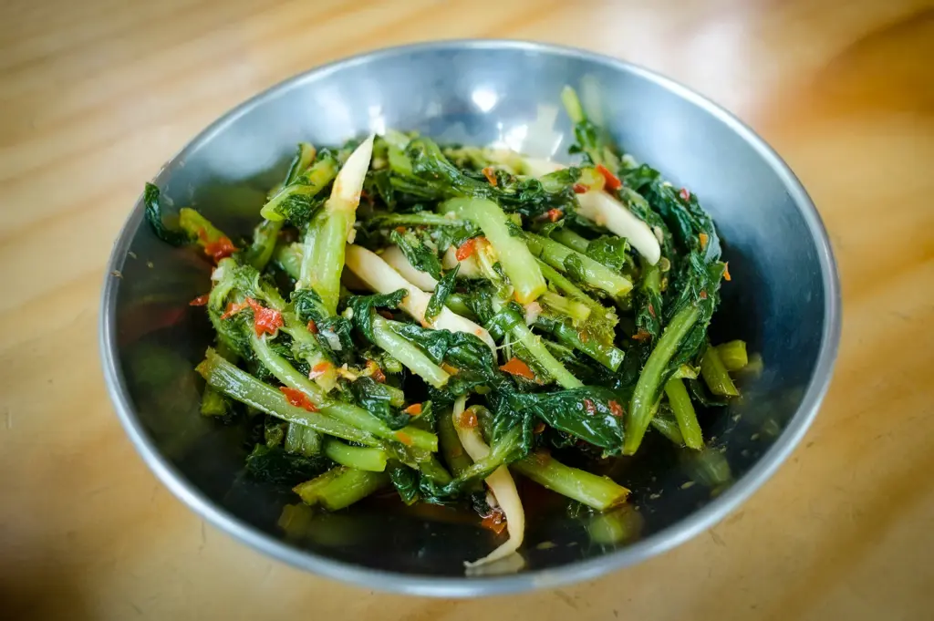 Spicy stir-fried leafy green vegetables with chili served in a metal bowl on a wooden table in Singapore