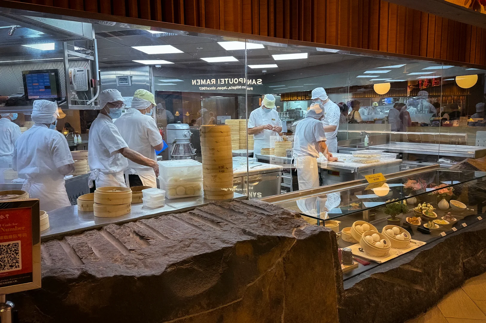 Open kitchen interior at Din Tai Fung Singapore with chefs preparing handmade dumplings and xiao long bao behind a glass viewing window beside dim sum display counters