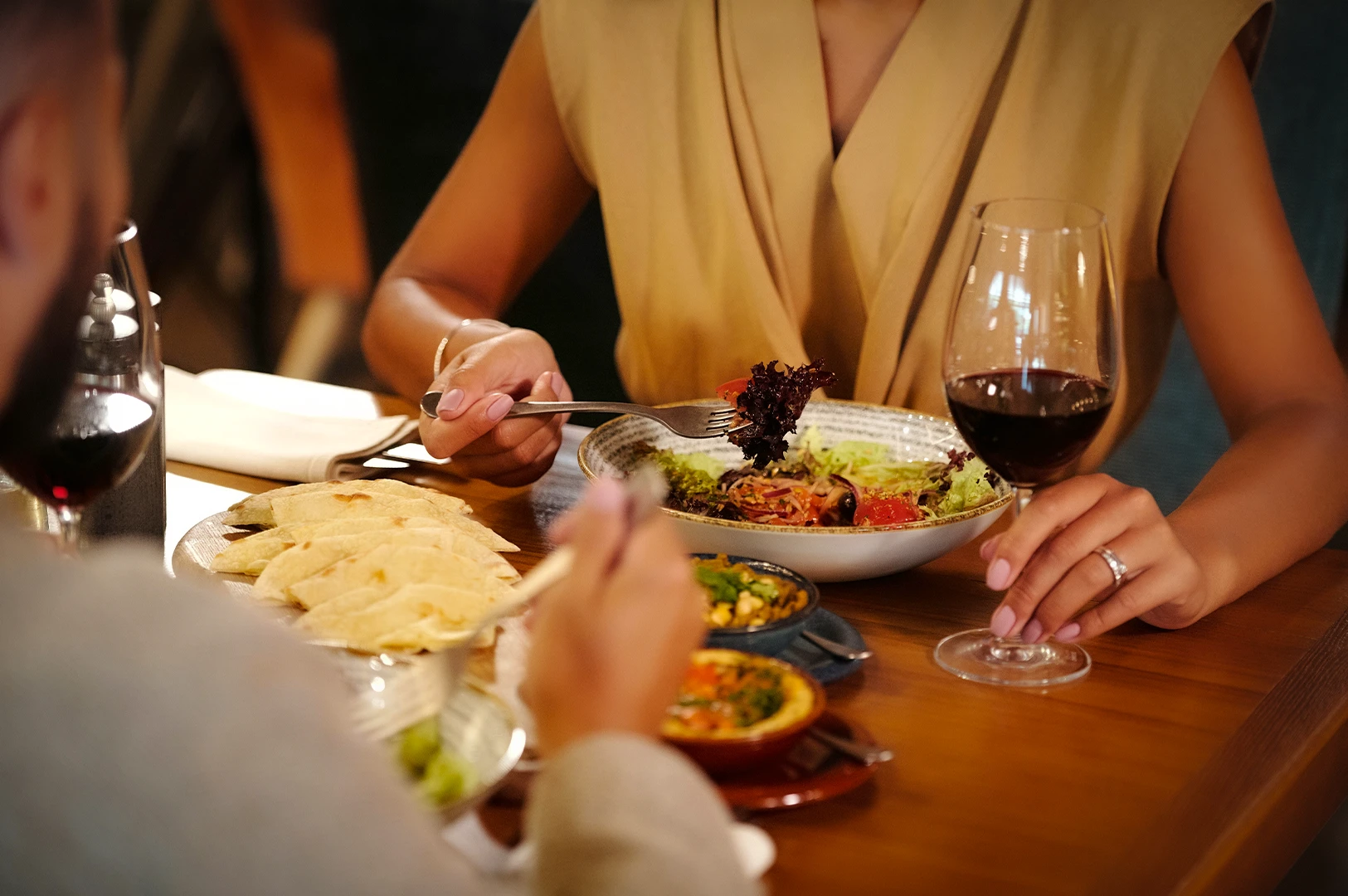 Couple enjoying a fresh salad at a restaurant table with flatbread, small dishes, and a glass of red wine.
