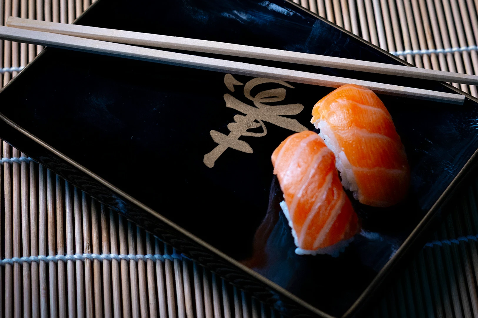 Two pieces of fresh salmon nigiri sushi served on a sleek black rectangular plate with a gold Japanese calligraphy character. Wooden chopsticks rest across the top of the plate, which is set on a traditional bamboo rolling mat.