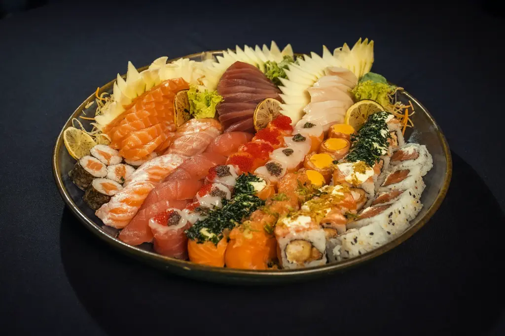 A large, colorful Japanese sushi and sashimi platter beautifully arranged on a dark plate. The assortment includes fresh slices of salmon and tuna sashimi, various nigiri, and a diverse selection of maki rolls topped with roe and garnishes. The platter is decorated with intricately carved ginger or radish fans and lemon slices.