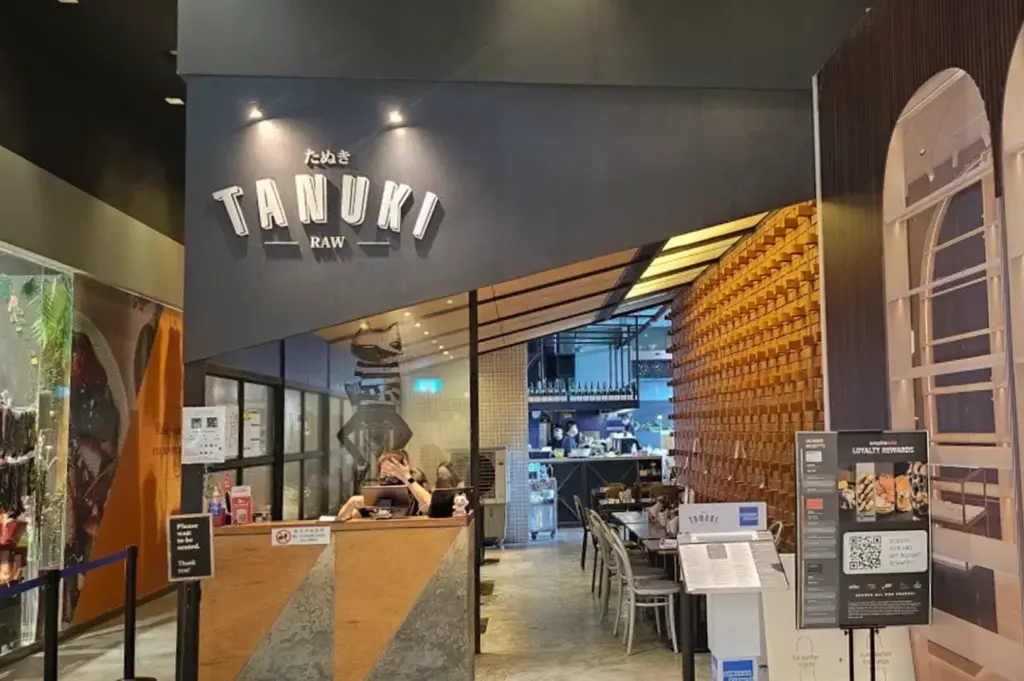 The storefront of Tanuki Raw in Singapore, featuring its modern industrial design with a prominent "TANUKI RAW" sign on a dark angled facade. The entrance shows a glimpse of the interior dining area with warm wood-textured walls, a bar counter in the back, and a host stand at the front.
