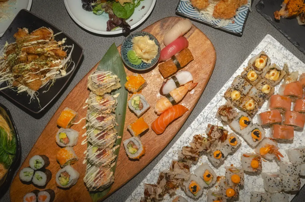 An extensive Japanese food spread featuring a variety of sushi and rolls on a textured tabletop. A wooden platter holds nigiri, including salmon, shrimp, and tuna, alongside maki rolls. Nearby plates display tempura, California rolls with roe, and specialty uramaki topped with bonito flakes and creamy sauces.