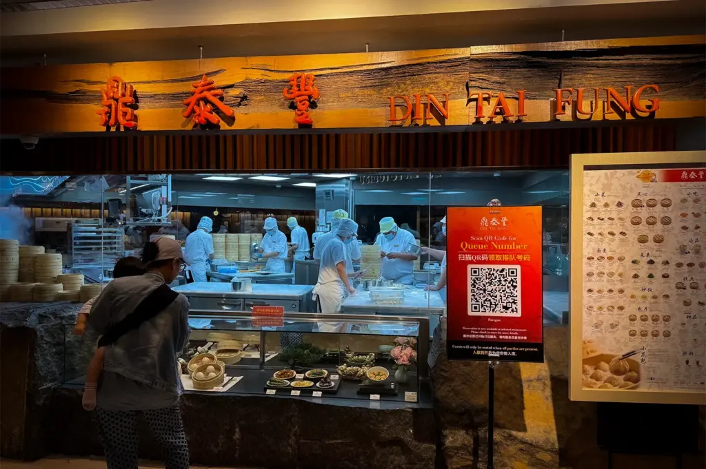 Din Tai Fung Singapore restaurant storefront featuring an open kitchen with chefs preparing dumplings, illuminated signage, menu displays, and customers viewing the handmade dim sum preparation area