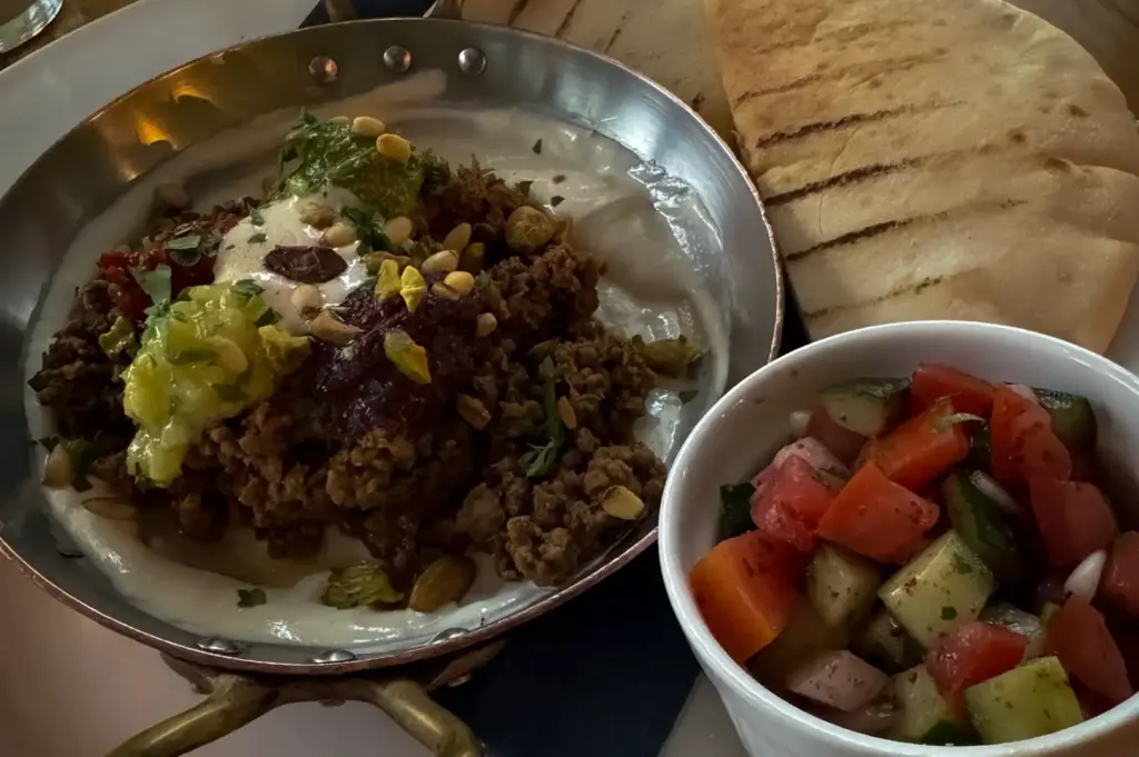 Middle Eastern style breakfast at Wild Honey Mandarin Singapore featuring minced lamb over creamy hummus, grilled pita bread, fresh cucumber tomato salad and iced coffee on a wooden dining table