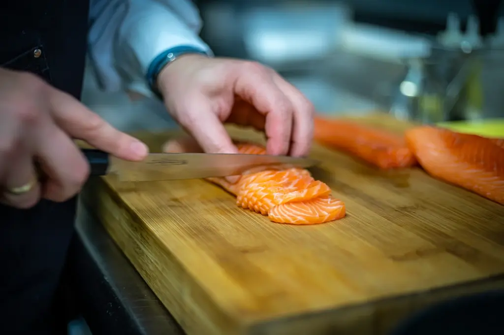 Close-up of a sushi chef’s hands skillfully slicing fresh, marbled salmon into thin sashimi pieces on a wooden cutting board. The chef uses a professional sharp knife to create precise cuts, with additional fillets ready in the background.