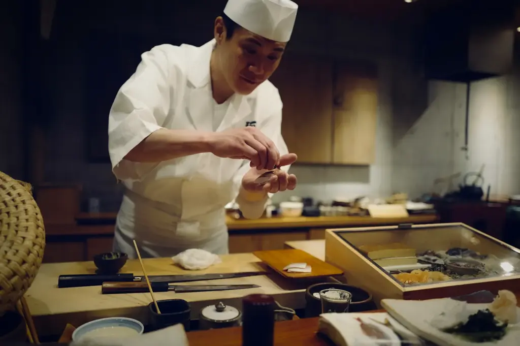 A focused Japanese sushi chef in traditional white attire and a chef's hat meticulously assembling nigiri behind a wooden counter. Professional sushi knives and a glass-topped ingredient box filled with fresh seafood are visible in the foreground of the authentic, dimly lit omakase-style restaurant.