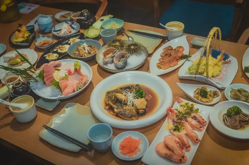 A lavish Japanese banquet spread across a light wooden table, featuring an assortment of authentic dishes. The meal includes a sashimi platter with fresh salmon and tuna on ice, sea urchin (uni), crispy shrimp tempura, grilled prawns, and a simmered fish head in soy-based broth. Small side dishes, condiments, and traditional ceramics complete the dining experience.