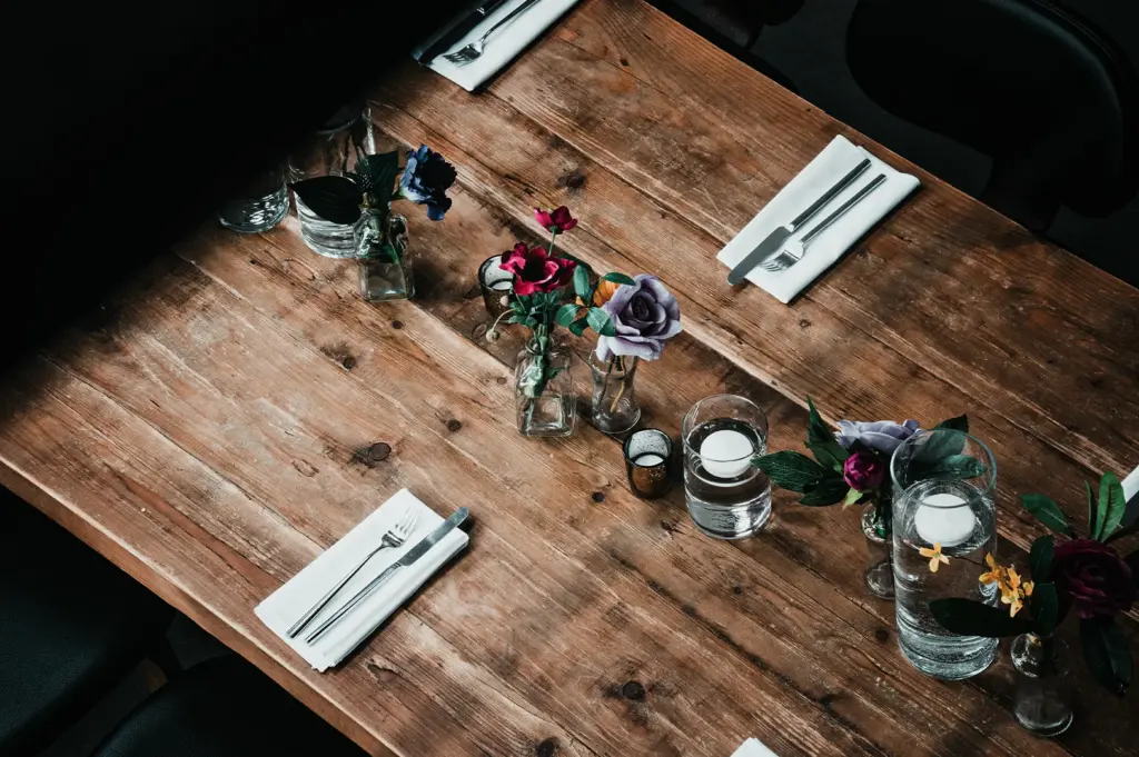 A high-angle view of a long, rustic wooden dining table elegantly set with white napkins, silverware, small glass vases holding colorful faux roses, and glass cylinders with floating candles.