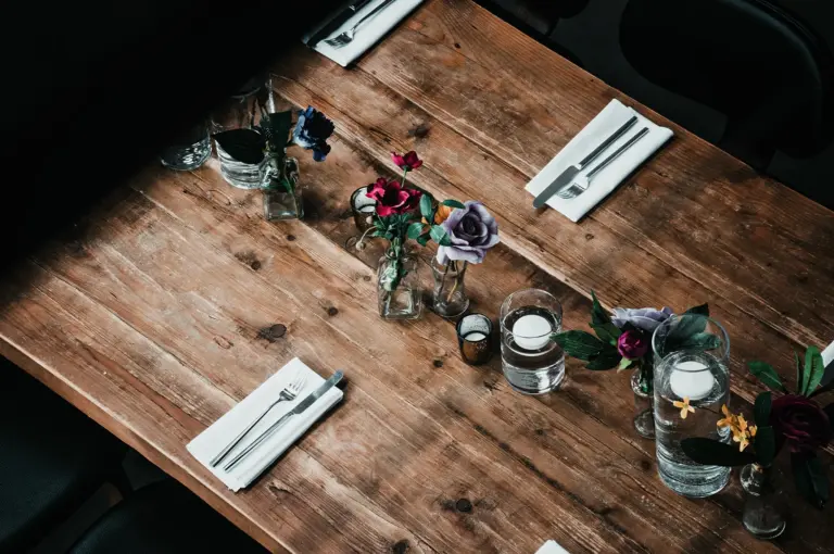 A high-angle view of a long, rustic wooden dining table elegantly set with white napkins, silverware, small glass vases holding colorful faux roses, and glass cylinders with floating candles.