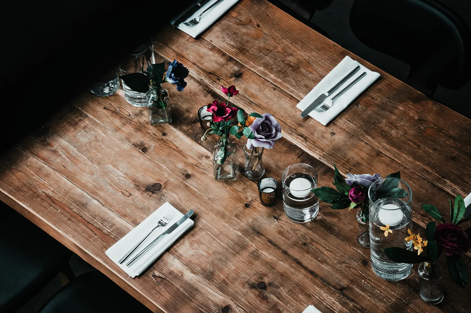 A high-angle view of a long, rustic wooden dining table elegantly set with white napkins, silverware, small glass vases holding colorful faux roses, and glass cylinders with floating candles.