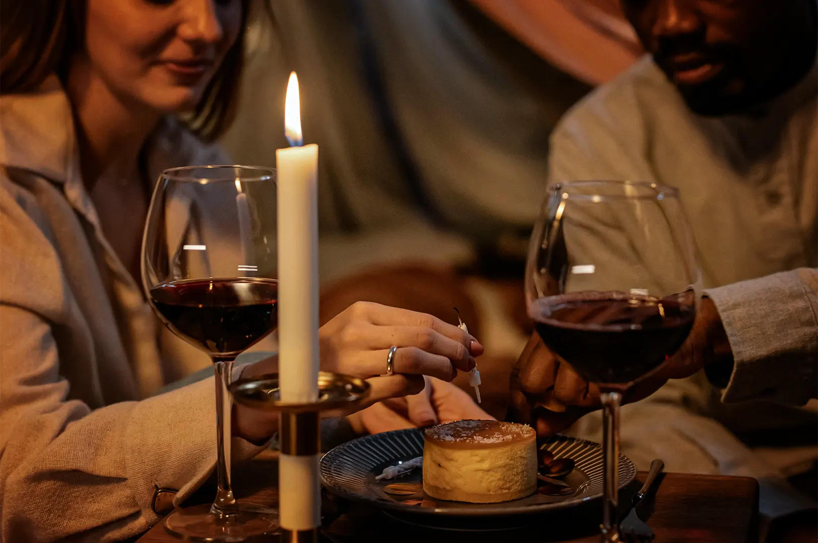 A warm, dimly lit medium close-up of a couple sharing an intimate moment at a table. In the center foreground, a small round cheesecake or flan sits on a dark plate, flanked by two large glasses of red wine. A single tall, white candlestick is lit on the left, casting a soft glow. In the blurred background, a woman on the left with a gentle smile and a man on the right are leaning in, their hands meeting near the dessert as the woman places a small unlit birthday candle onto the cake.