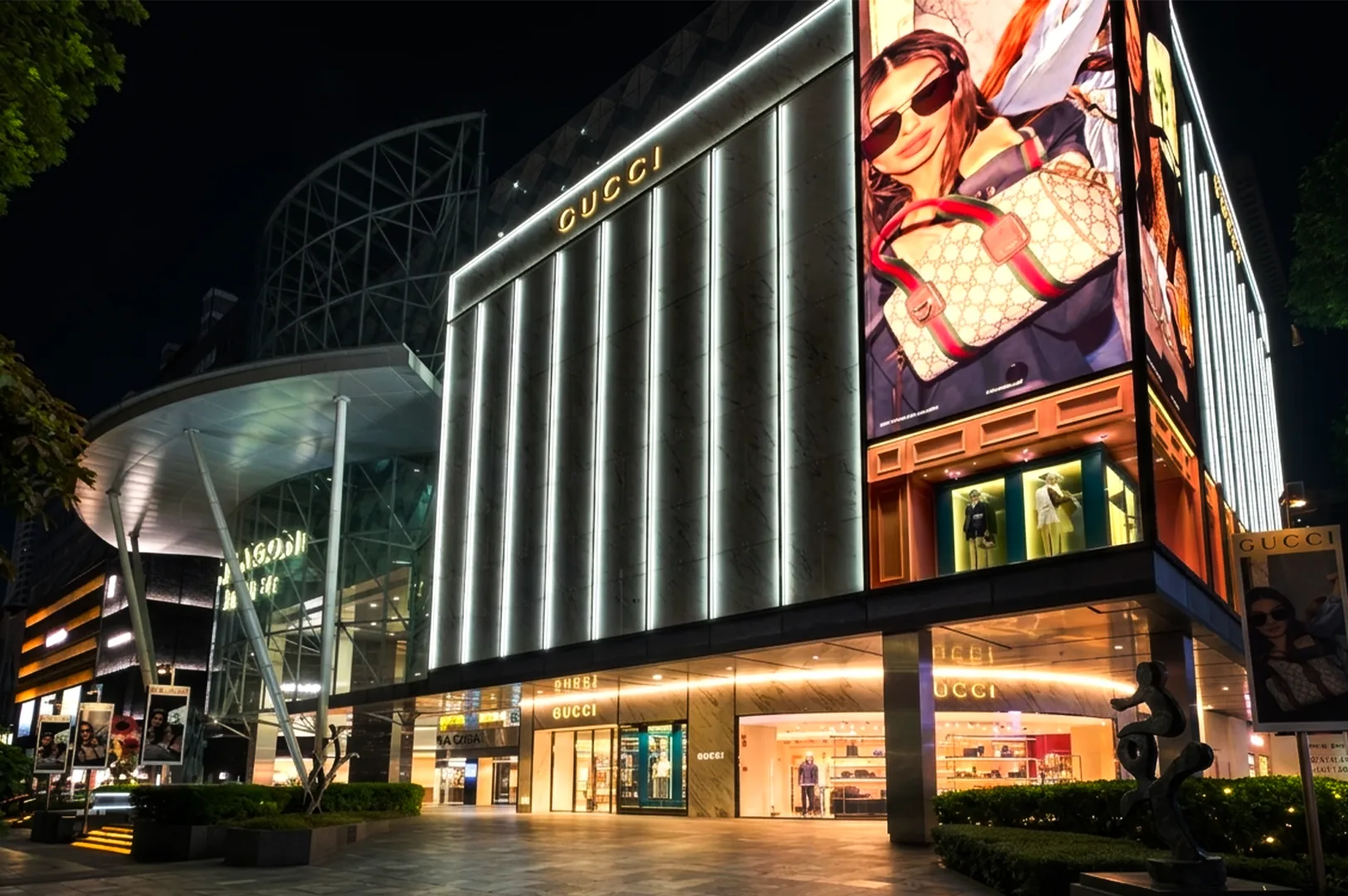 A night shot of the luxury Gucci storefront on Orchard Road in Singapore, featuring a modern marble facade illuminated by vertical LED light strips, an oversized digital billboard showing a fashion model, and bright window displays.