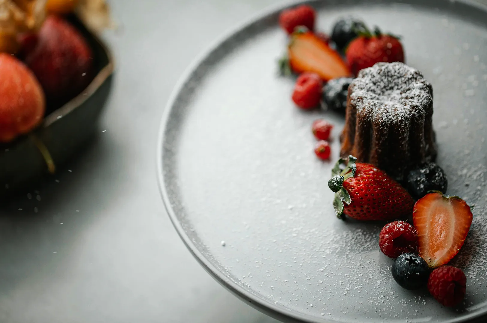 A small, fluted chocolate cake or canelé dusted with powdered sugar, elegantly arranged on a minimalist grey plate alongside an assortment of fresh strawberries, raspberries, and blueberries.