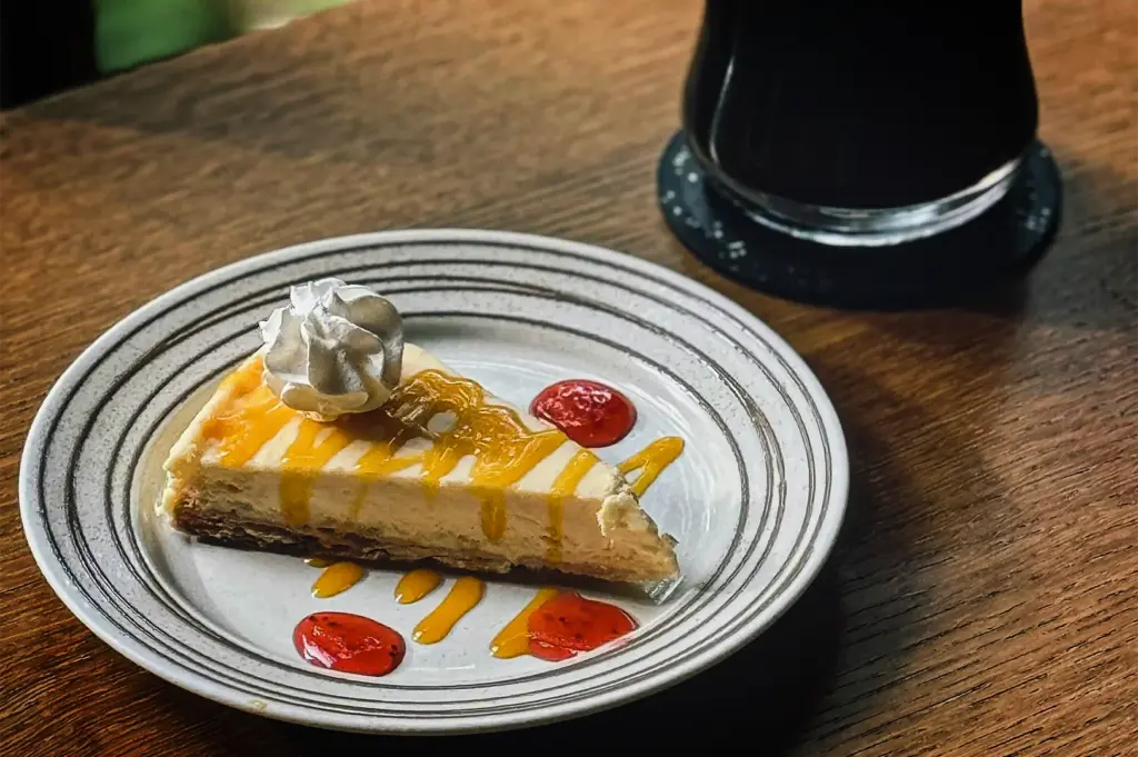 A slice of cheesecake sits on a white plate with concentric grey rings, placed on a wooden table. The cheesecake has a dark crust, a creamy filling, and is drizzled with a bright yellow mango or passionfruit sauce, topped with a dollop of whipped cream. Three pools of red strawberry gel surround the slice on the plate. In the blurred background, a dark drink in a glass rests on a black coaster.