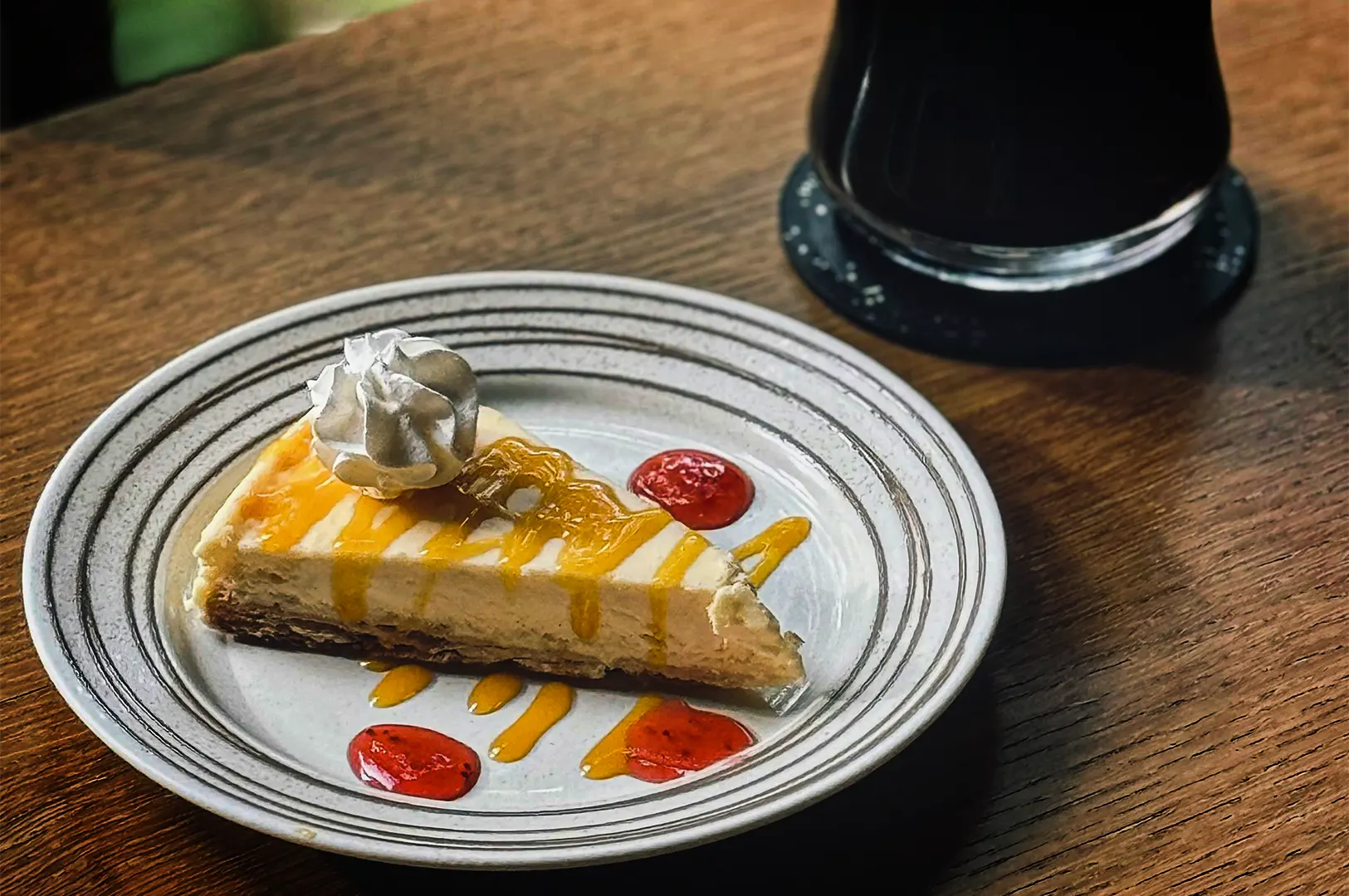 A slice of cheesecake sits on a white plate with concentric grey rings, placed on a wooden table. The cheesecake has a dark crust, a creamy filling, and is drizzled with a bright yellow mango or passionfruit sauce, topped with a dollop of whipped cream. Three pools of red strawberry gel surround the slice on the plate. In the blurred background, a dark drink in a glass rests on a black coaster.
