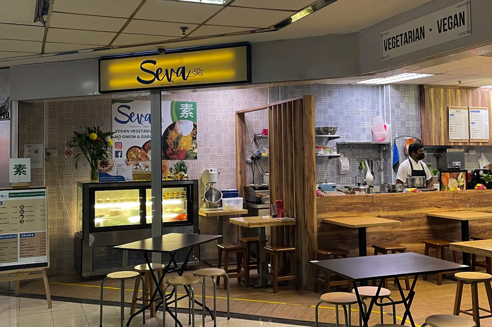 A small vegetarian and vegan restaurant storefront inside a food center, featuring wooden tables and stools, a warmly lit counter, and a display case with food items. A staff member works behind the open kitchen area, while signage above the entrance highlights the restaurant’s vegetarian and vegan offerings in a casual and welcoming dining setting.