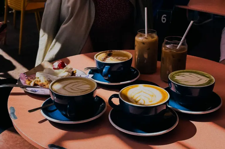 A round pink outdoor café table heavily laden with drinks, including four hot lattes in dark blue ceramic cups showcasing intricate latte art, two tall glasses of iced coffee with straws, and a small plate with a slice of cake topped with cream and a strawberry.