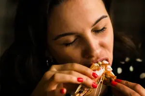 A close-up portrait of a woman with her eyes closed, completely focused as she takes a bite out of a large, freshly cooked shrimp or prawn that she is holding with both hands.
