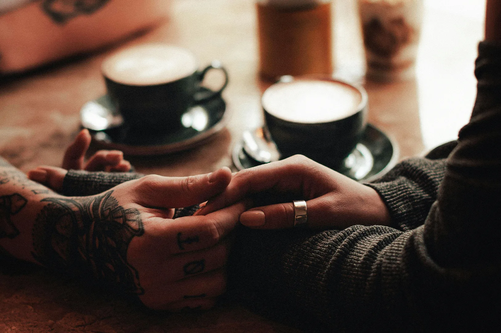 Close-up of two people holding hands across a table in a cozy café, with two cups of coffee in the blurred background. One person wears a thick sweater and a silver ring, while the other has detailed tattoos on their hand and wrist.