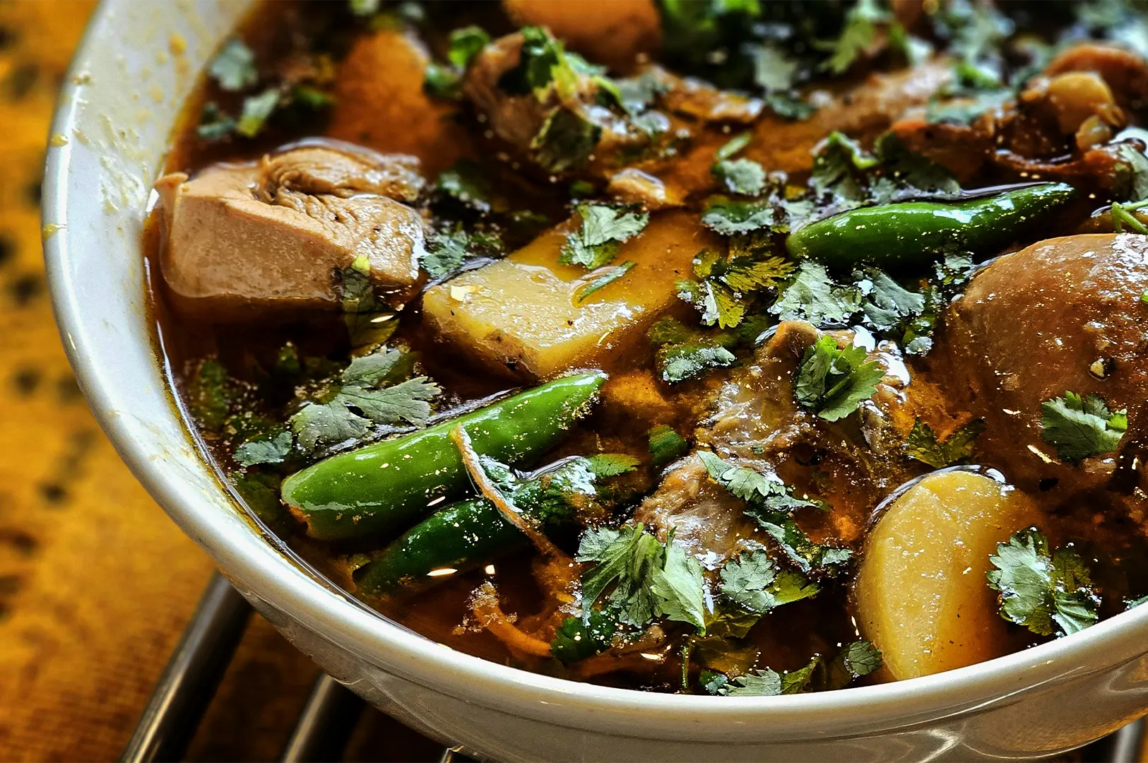 A close-up, high-angle shot captures a rich and deeply aromatic bowl of soup curry, representing the file "1-Soup Curry The Bowl That Rewards You.webp". Served in a white ceramic bowl resting over a metallic wire stand, the dish features a dark, glossy, and complex spice-infused broth. It is packed with hearty ingredients, including tender chunks of braised chicken meat, soft-stewed potatoes, and whole bright green chili peppers. A generous, vibrant garnish of freshly chopped cilantro leaves is scattered across the entire surface, catching the light and completing this warm, comforting culinary presentation.