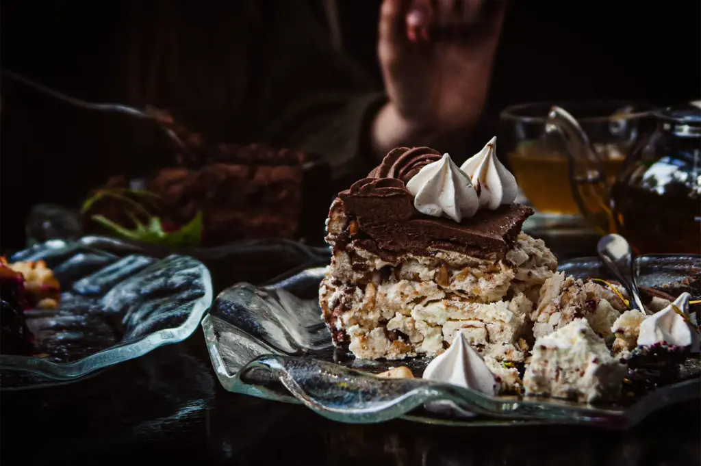 A moody, close-up shot of a large slice of Dacquoise or layered meringue cake resting on a wavy, clear glass plate. The cake features rich layers of crushed nuts, light cream, and a thick chocolate fudge topping decorated with swirls of chocolate frosting and white piped meringue kisses. In the softly blurred background, a person’s hand is visible alongside a dark chocolate dessert and a warm glass teapot.