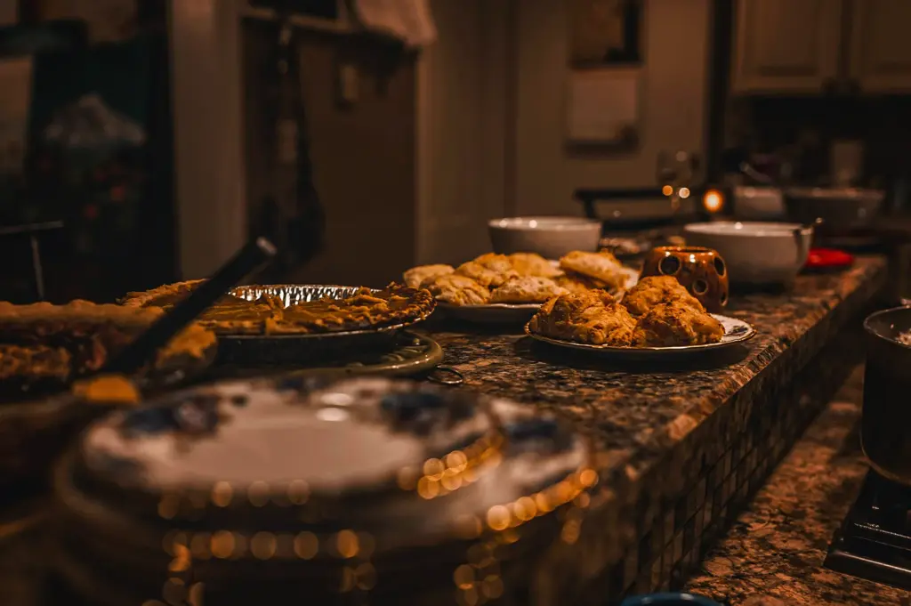A long, granite kitchen island counter serving as a buffet line under warm, dim lighting, laden with sweet and savory dishes including whole pies, fresh baked scones or biscuits, cookies, and serving bowls.