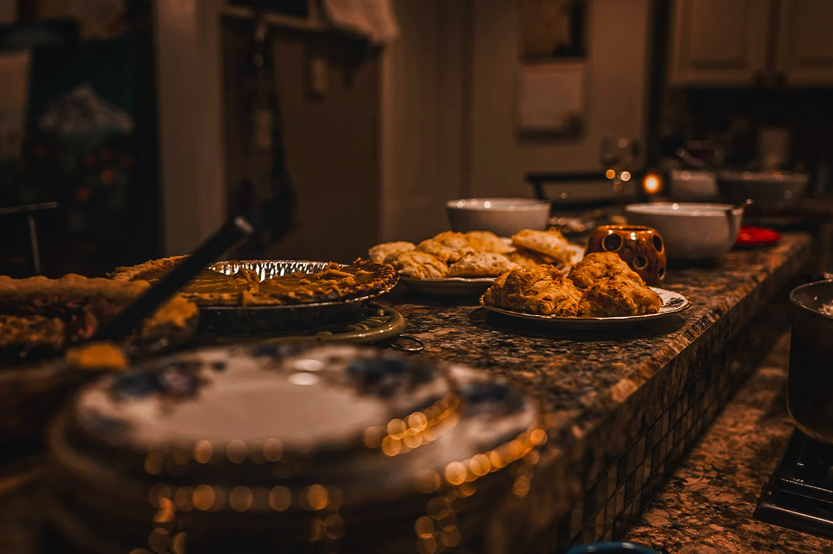 A long, granite kitchen island counter serving as a buffet line under warm, dim lighting, laden with sweet and savory dishes including whole pies, fresh baked scones or biscuits, cookies, and serving bowls.