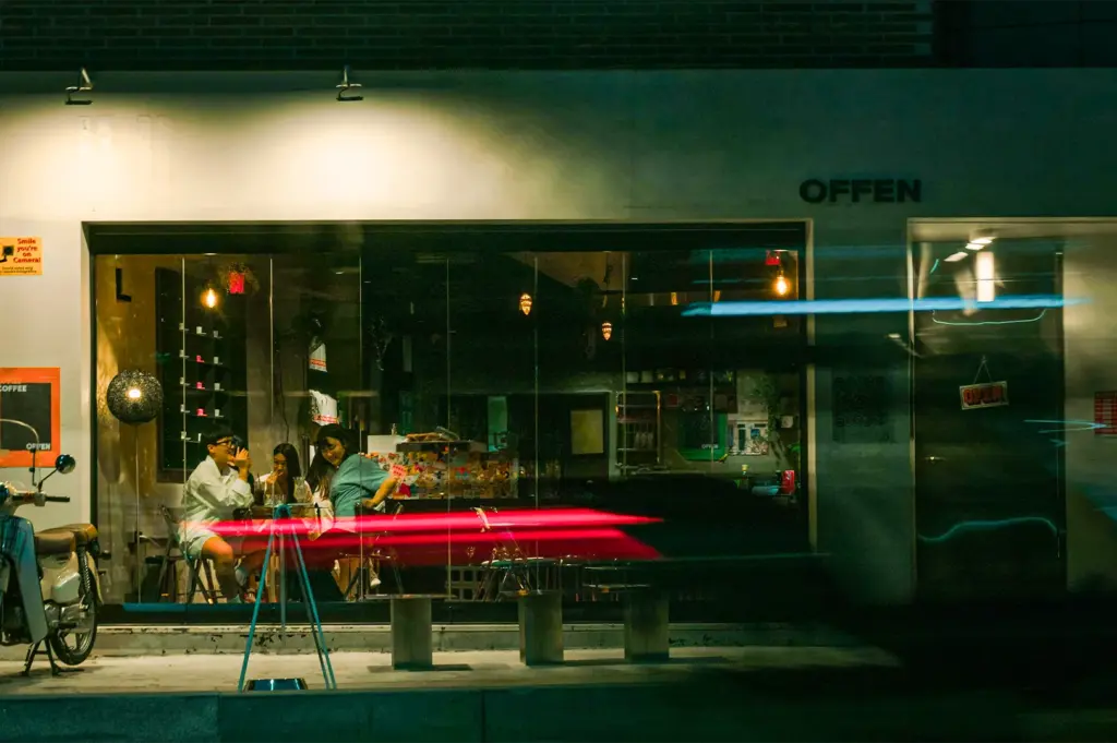A nighttime view of a modern café storefront named "OFFEN" with a large glass window showing patrons inside, contrasted by long-exposure light streaks from passing traffic in the foreground.