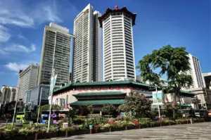 A low-angle, wide shot captures the iconic architectural fusion of Tang Plaza along a bustling city street, representing the file "1-The Five Best Dessert Shops in Orchard.webp". In the center stands a prominent octagonal white tower topped with a traditional red Chinese pagoda-style roof, flanked by several modern, sleek skyscrapers under a vibrant blue sky with wispy clouds. Below the towers, the main complex features multi-tiered green-tiled roofs decorated with red and white banners along the facade. Lush, manicured green shrubbery, blooming flower beds, and mature trees line the wide, patterned stone sidewalk in the foreground, where city buses and street lamps frame the lively urban scene.