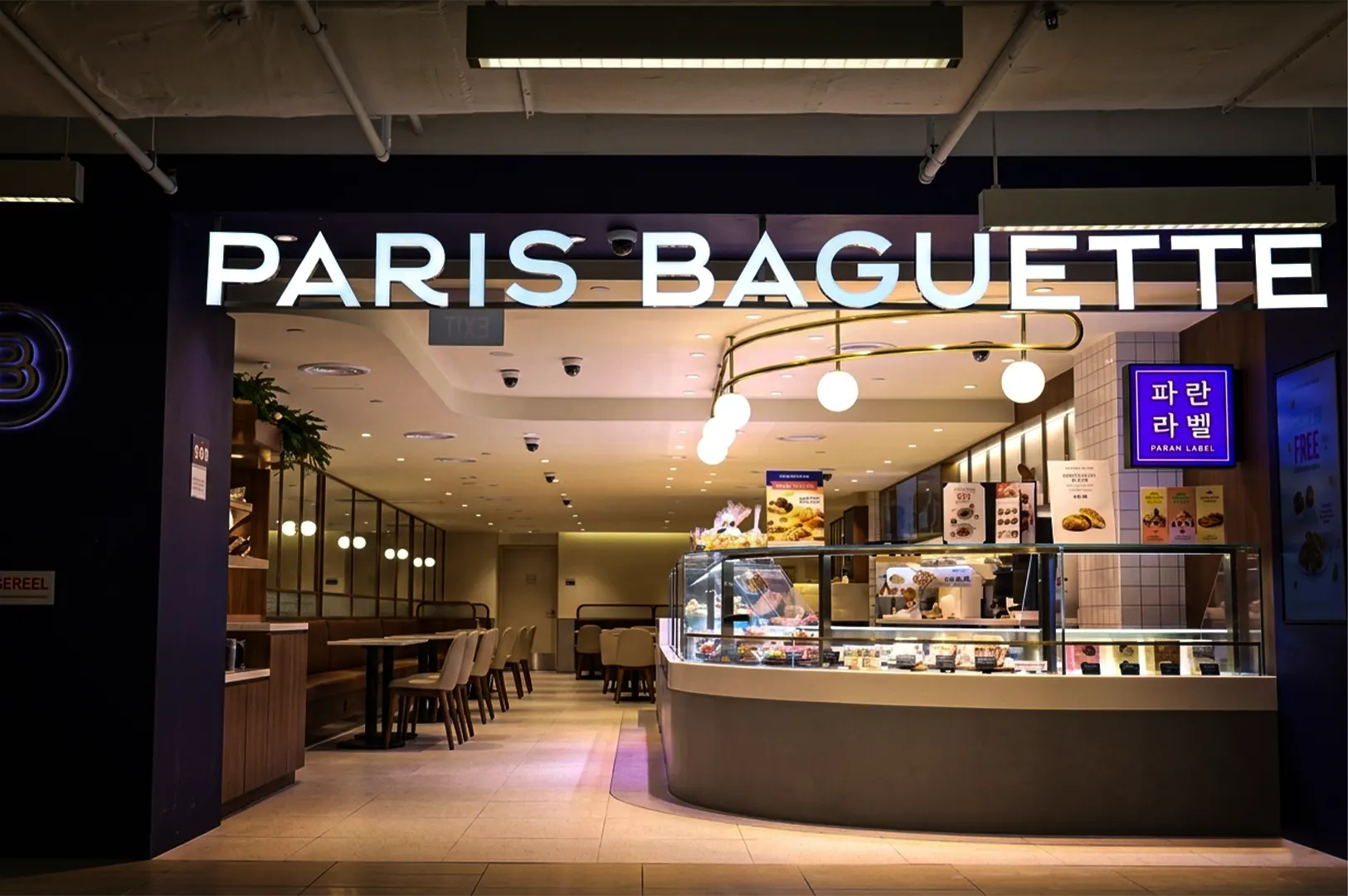 An eye-level shot of the entrance to a modern, brightly lit "PARIS BAGUETTE" bakery and café. The prominent white storefront sign glows above a curved glass display counter on the right filled with pastries. To the left, a long seating area features booth benches and small dining tables. The interior has warm, minimalist decor with light tiled floors, neutral-colored chairs, and a curved brass light fixture holding glowing globe bulbs over the main counter. A small blue sign with Korean text hangs on the right wall.