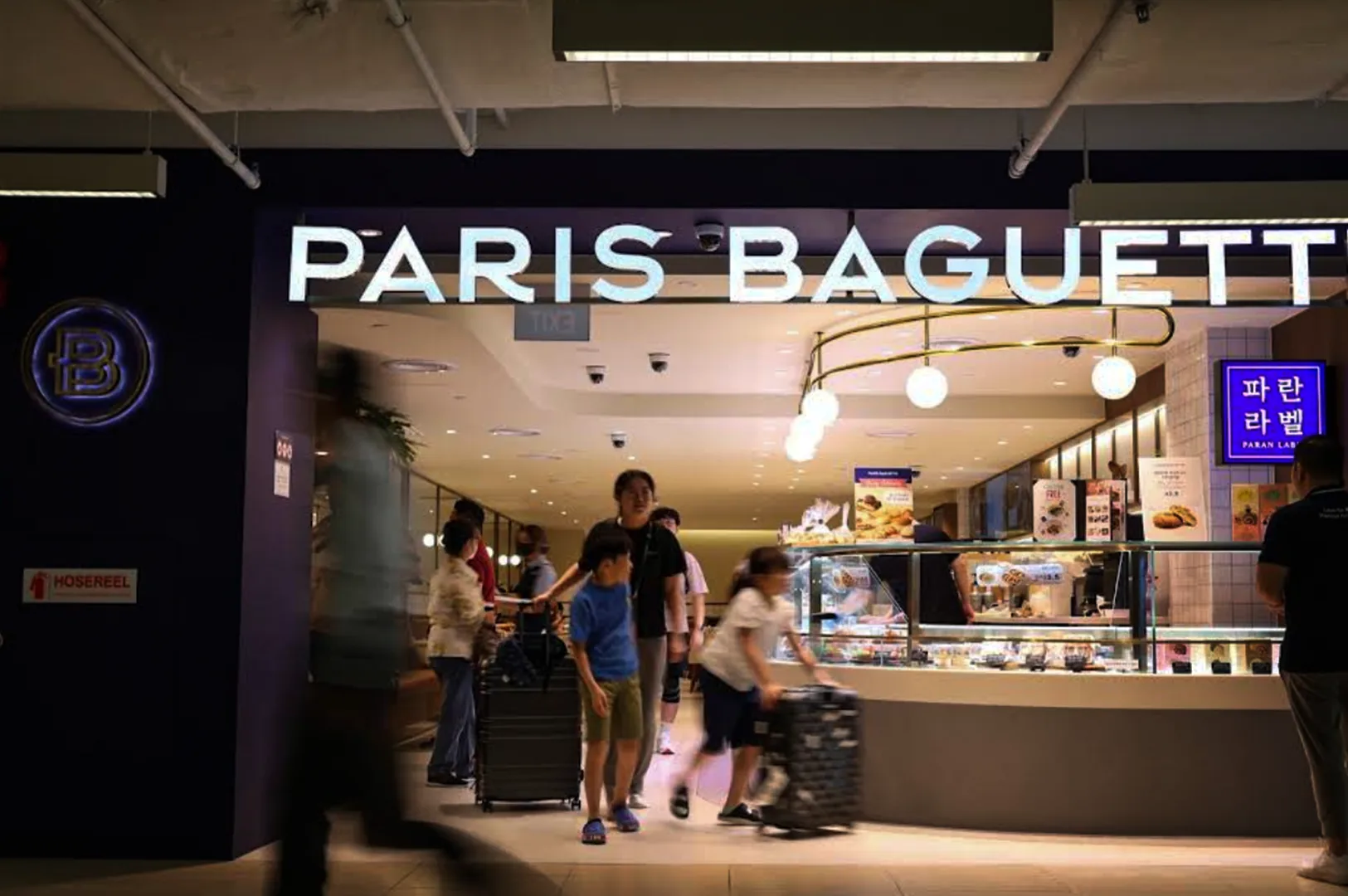 A bright storefront of a "PARIS BAGUETTE" bakery located inside a modern commercial building or mall, featuring an illuminated white sign, glass display cases filled with pastries, and several patrons with luggage walking past the entrance.