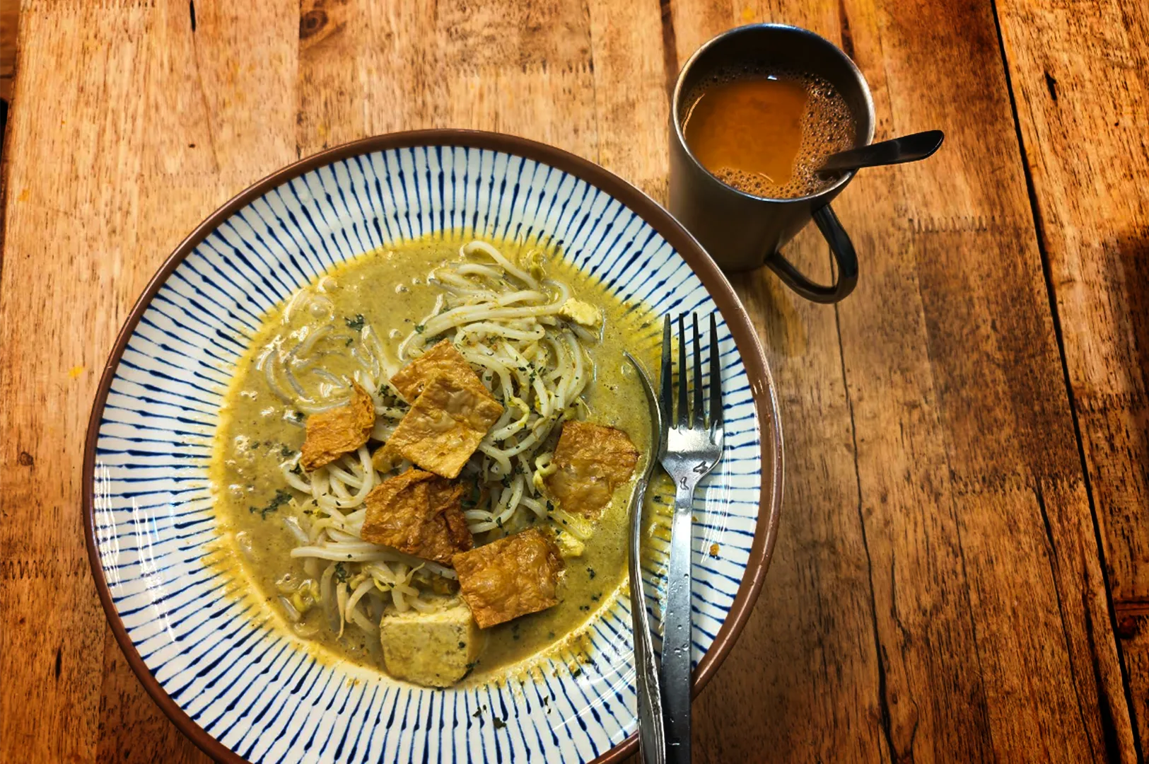 A top-down view of a bowl of noodles in creamy curry broth topped with crispy tofu pieces, served on a patterned ceramic plate atop a rustic wooden table. A fork rests beside the noodles, while a mug of hot tea or coffee sits nearby, creating a warm and comforting meal setting.