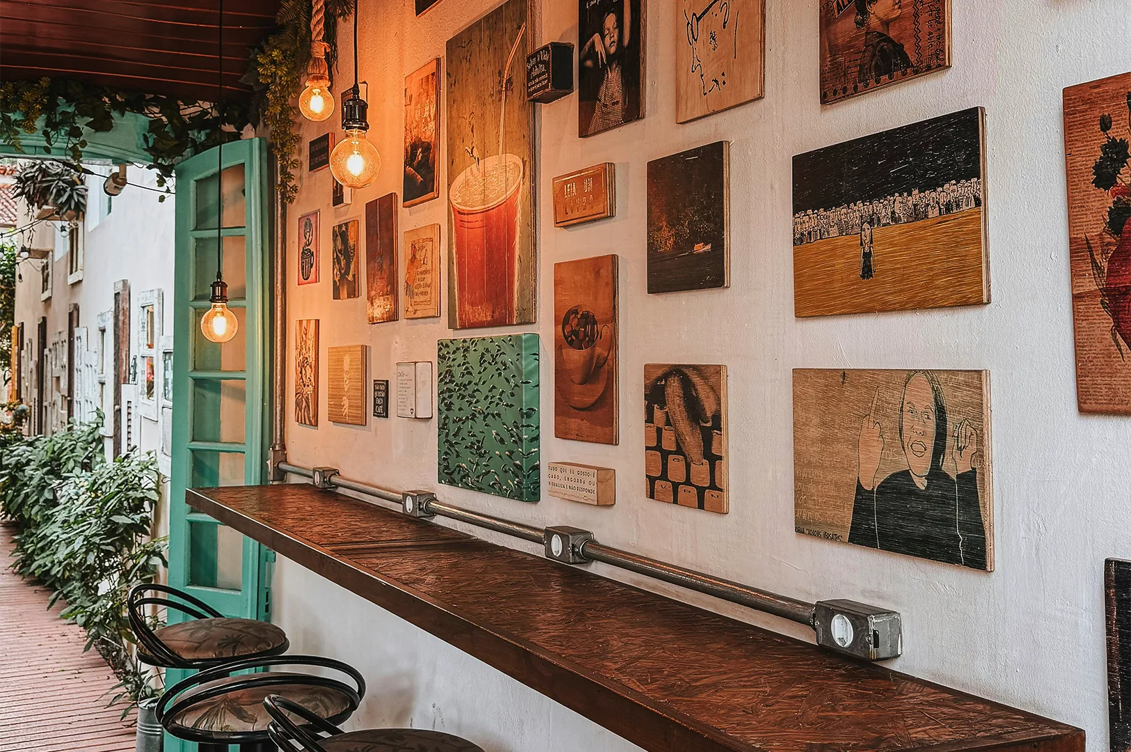 A long, dark wooden bar counter lined with bar stools inside a trendy, narrow café. The white focal wall is covered in an eclectic gallery of wooden art plaques and paintings, illuminated by warm, glowing Edison bulb pendant lights.