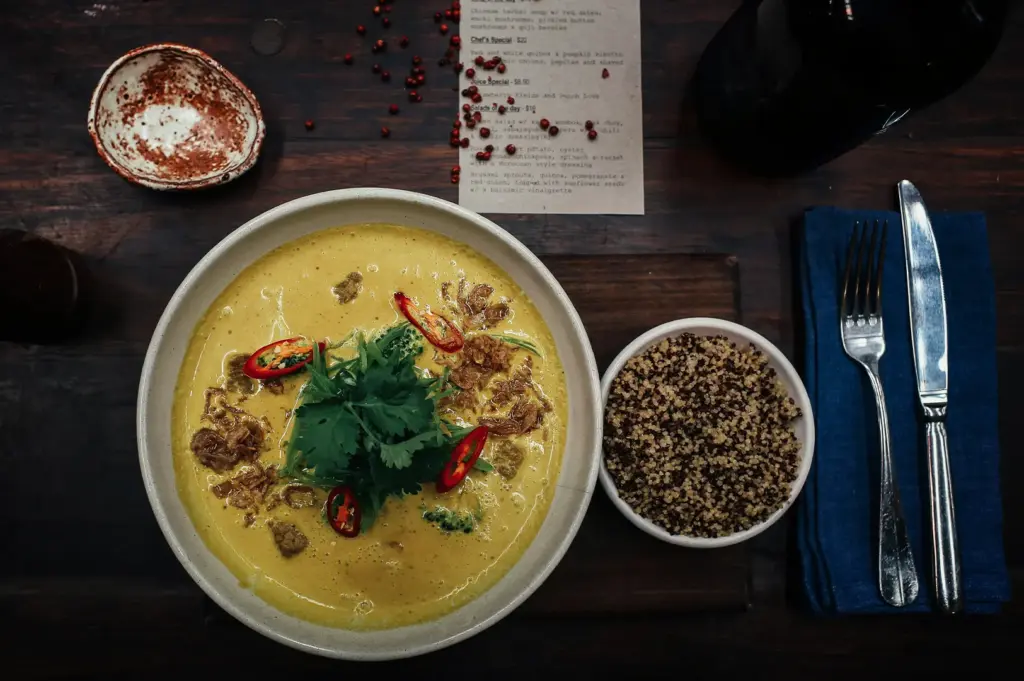 A detailed overhead shot captures an elegant flat-lay arrangement of a gourmet meal on a dark, rustic wooden table, representing the file "2-Soup Curry The Bowl That Rewards You.webp". The main attraction is a large, off-white ceramic bowl filled with a creamy, bright yellow curry soup garnished with a dense bunch of fresh green cilantro in the center, sliced red chilies, broccoli florets, and crispy fried shallots. To its right sits a smaller white bowl filled with a blend of cooked white and red quinoa, positioned next to a blue fabric napkin topped with a silver fork and knife. Completing the restaurant scene in the upper portion of the frame are a small textured spice dish, a scattering of dried pink peppercorns, a printed paper menu, and the edge of a dark glass bottle, all adding to the moody and sophisticated dining atmosphere.