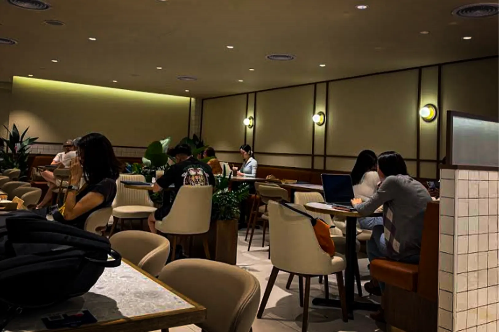 An alt text description for the image 2-Why Paris Baguette Remains.webp: An eye-level shot of a dimly lit, cozy café dining room with several patrons seated at small round tables. People are seen working on laptops, reading, or conversing in cushioned, cream-colored armchairs. The space features warm ambient lighting from recessed ceiling lights and a row of glowing spherical wall sconces mounted on wood-paneled walls. Large green potted plants are placed between tables, adding a touch of nature to the relaxed atmosphere.
